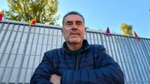 Un uomo in primo piano davanti a una recinzione con bandiere sullo sfondo