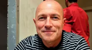 Un uomo seduto in un interno, sorride mentre un'altra persona è visibile sullo sfondo.