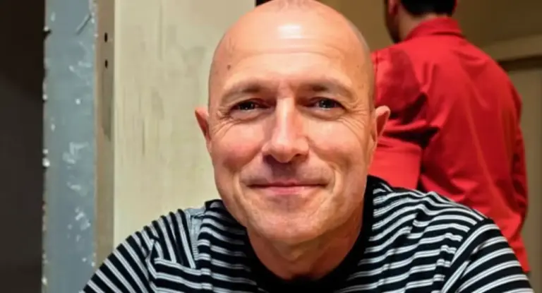 Un uomo seduto in un interno, sorride mentre un'altra persona è visibile sullo sfondo.