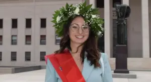 Una persona con corona d'alloro e fascia rossa posa davanti a un edificio universitario