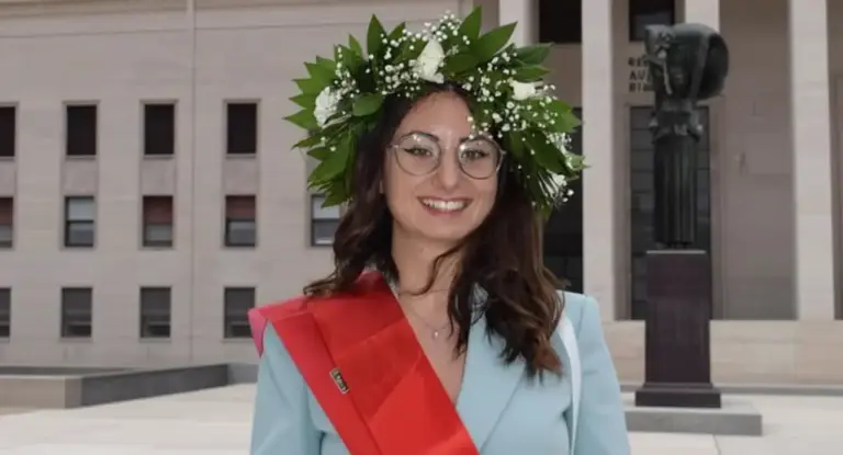 Una persona con corona d'alloro e fascia rossa posa davanti a un edificio universitario