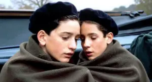 Due bambini avvolti in una coperta, con espressioni serie, indossano berretti scuri