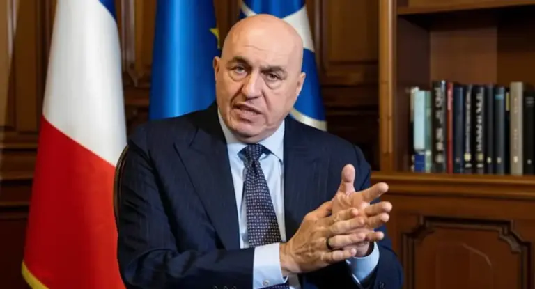 Un uomo in abito formale è seduto davanti a bandiere e uno scaffale di libri