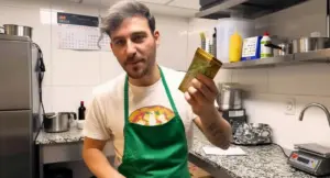 Un pizzaiolo con grembiule verde mostra una banconota all'interno di una cucina professionale.