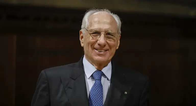 Una persona anziana in abito formale sorride davanti a uno sfondo scuro