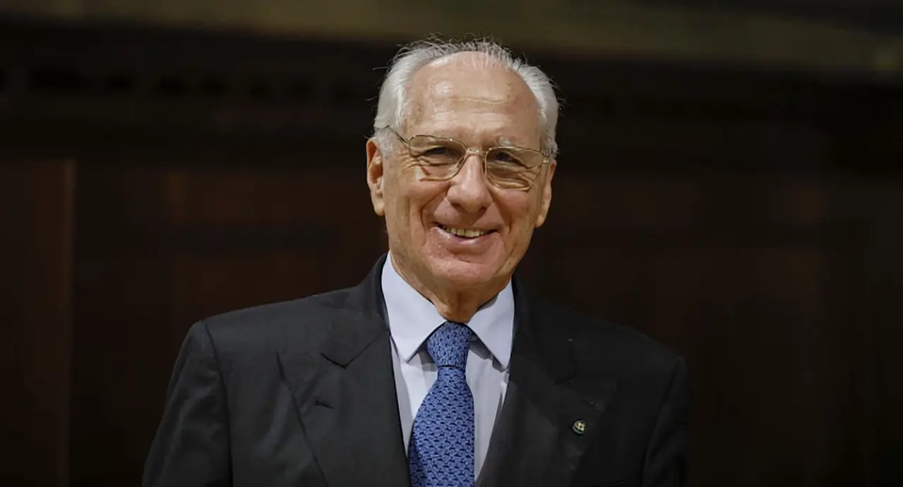 Una persona anziana in abito formale sorride davanti a uno sfondo scuro
