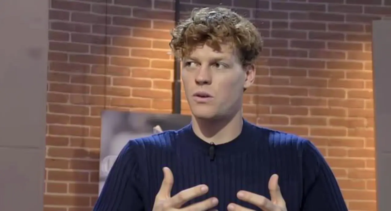 Un uomo con maglione blu parla durante un'intervista in un ambiente interno con sfondo a mattoni